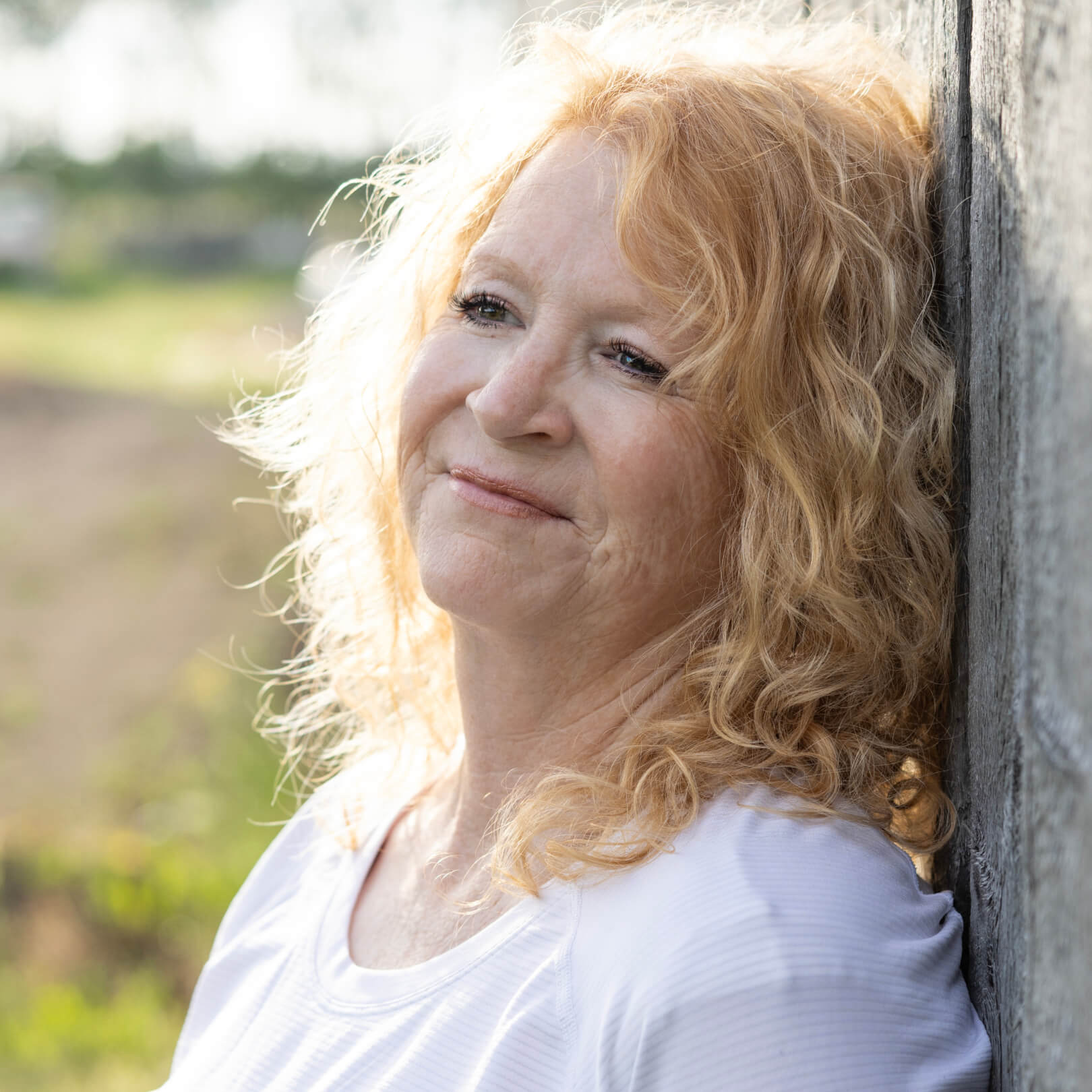 A woman with curly blonde hair leans against a wall, smiling softly in an outdoor setting.