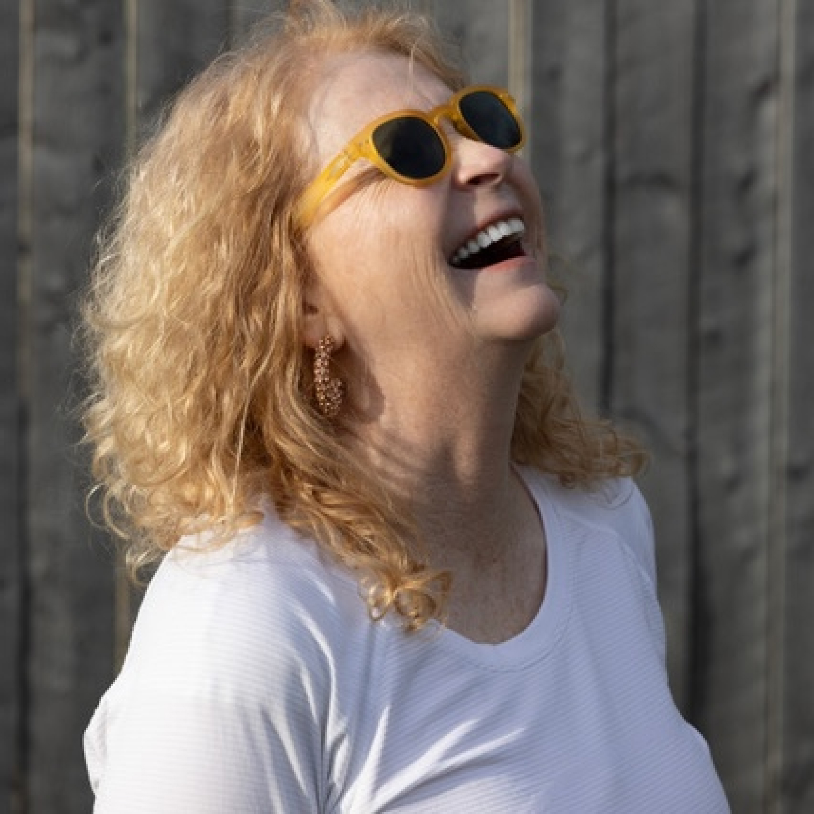 A woman with curly blonde hair and yellow sunglasses laughs, looking upward. She wears a white shirt. The background is a wooden fence.